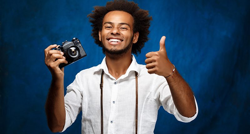 Young handsome african man holding old camera over blue background.