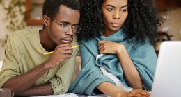 Young African wife and husband with many debts doing paperwork and tax returns together, analyzing expenses, planning family budget and calculating bills, sitting at kitchen table with laptop, calculator and papers